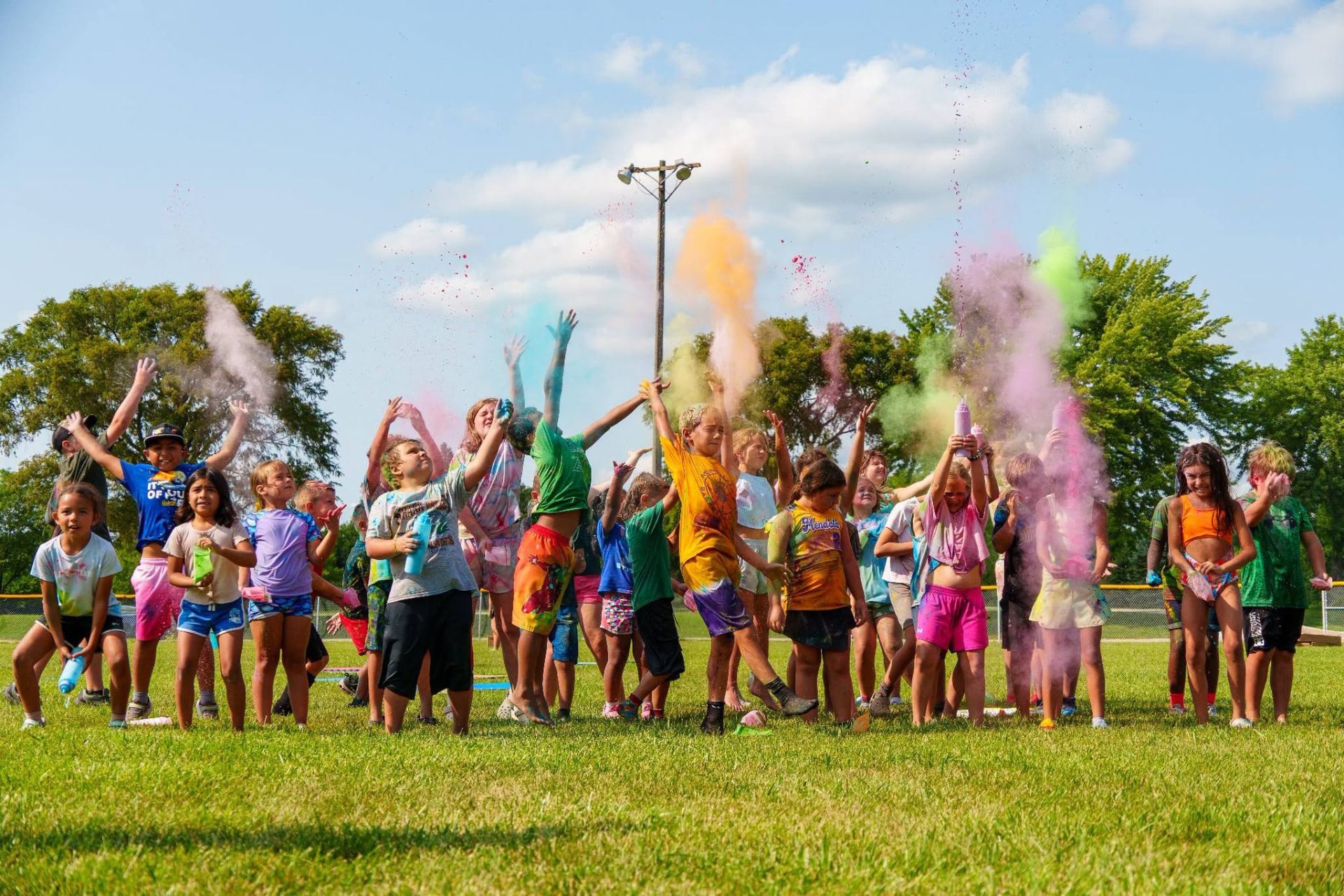 Summer camp kids tossing color powder into the air together on a sunny ball field